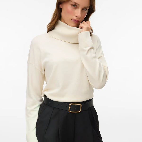 Women's birch colored turtleneck sweater with long sleeves and relaxed fit styled with tailored brown trousers.