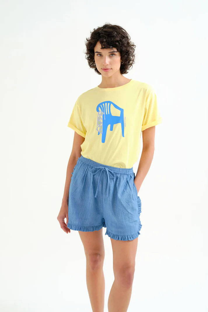 Women's pastel yellow graphic T-shirt featuring a bold blue chair illustration, rolled sleeves, crew neckline, and relaxed fit for casual everyday style.