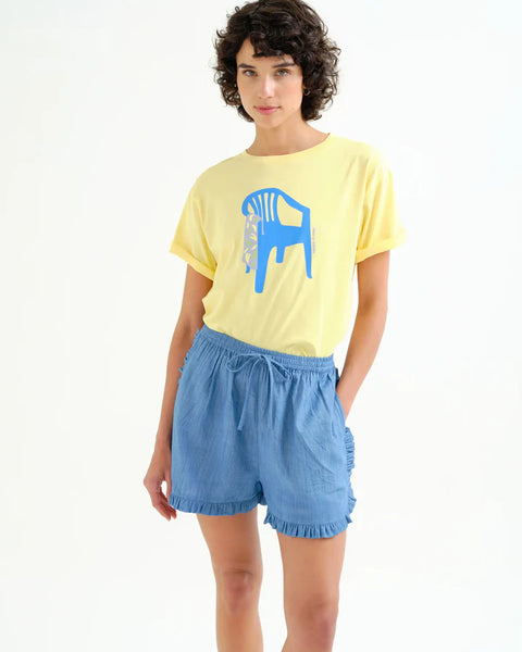 Women's pastel yellow graphic T-shirt featuring a bold blue chair illustration, rolled sleeves, crew neckline, and relaxed fit for casual everyday style.